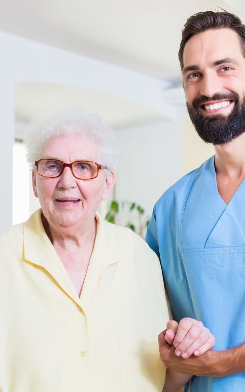 junge männliche Pflegekraft stützt eine ältere Dame, beide lächeln